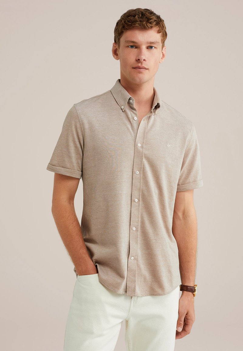 Giovane uomo dai capelli ricci e corti, vestito con una camicia beige a maniche corte con bottoni e pantaloni bianchi, in piedi con una mano in tasca.