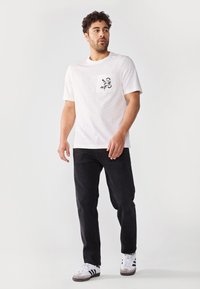 White short-sleeve t-shirt featuring a black floral pocket print, paired with dark jeans and white athletic shoes with black accents.