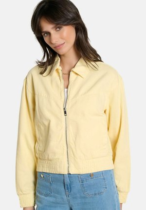 Frau trägt eine hellgelbe Jacke mit Reißverschluss, Kragen und elastischen Bündchen, kombiniert mit hellblauen Jeans mit Fronttaschen.