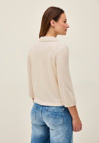 Beige, texturerad topp med krage och trekvartsärmar, kombinerad med ljusblå denimjeans med bakfickor.