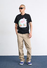 Maglietta nera con una grafica colorata di un fenicottero, abbinata a pantaloni beige e sneaker blu, su uno sfondo semplice.