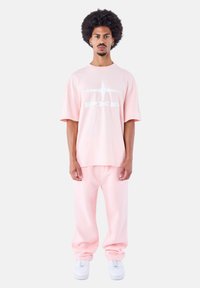 Camiseta oversized de color rosa claro con logotipo blanco, combinada con pantalones de chándal rosa a juego y zapatillas blancas, con un ajuste relajado y textura suave.