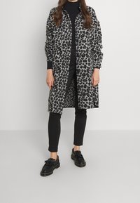 Grauer Leopardenmuster langer Mantel mit Knopfverschluss und Seitentaschen, kombiniert mit schwarzen, schmal geschnittenen Hosen und schwarzen Slippern. Glatte Textur.