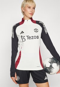 Fehér és fekete, hosszú ujjú futball edzőpóló piros részletekkel, Manchester United logóval és "Tezos" felirattal, fekete rövidnadrággal párosítva.