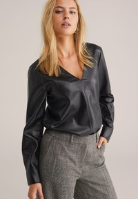 Schwarze Kunstlederbluse mit einem V-Ausschnitt und langen Ärmeln, kombiniert mit grauen karierten maßgeschneiderten Hosen. Glatte Textur, taillierte Silhouette.