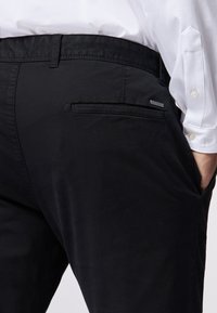 Schwarze Hose aus glattem Stoff mit flachem Bund, dezenten Taschen-details und einem kleinen Markenetikett auf der Rückseite.