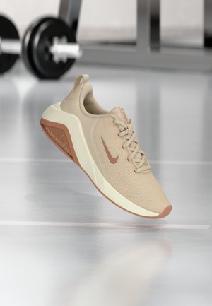 Chaussure de course Nike beige avec semelle et logo marron, suspendue au-dessus du sol de la salle de sport avec des haltères floutées en arrière-plan.