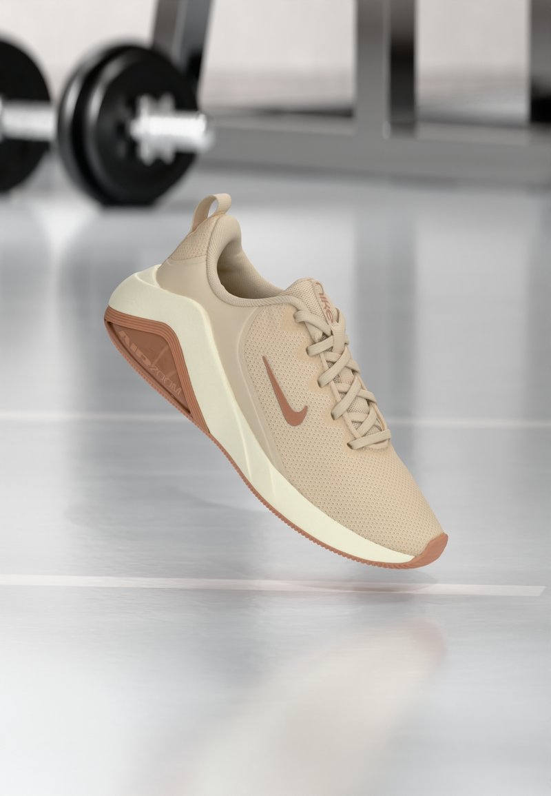Chaussure de course Nike beige avec semelle et logo marron, suspendue au-dessus du sol de la salle de sport avec des haltères floutées en arrière-plan.