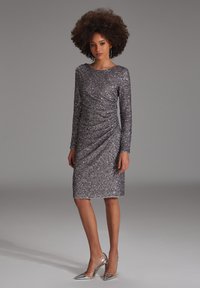 Robe en sequins argentés avec manches longues, décolleté en scoop et détails froncés. Associée à des escarpins métalliques à bout pointu.