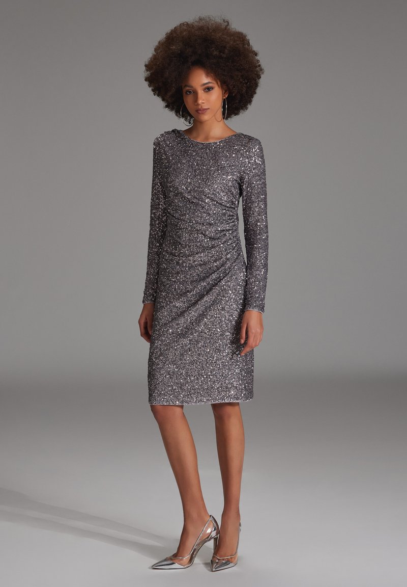 Robe en sequins argentés avec manches longues, décolleté en scoop et détails froncés. Associée à des escarpins métalliques à bout pointu.