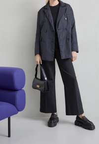 Veste noire à double boutonnage avec boutons, assortie à un haut noir et un pantalon large. Sac en cuir noir et chaussures noires imposantes.