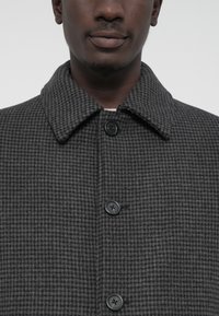 Férfi sötétszürke houndstooth mintájú kabátot visel, nagy gallérral és három látható fekete gombbal, amely megmutatja a nyakát és az alsó arcát.