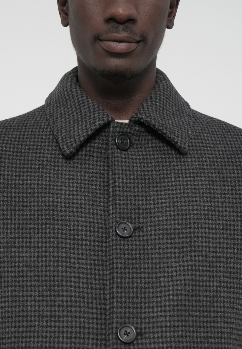 Homme portant un manteau à motif pied-de-poule gris foncé avec un grand col et trois boutons noirs visibles, montrant le cou et le bas du visage.