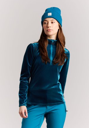 Vrouw met lang bruin haar die een blauwe muts draagt, een teal fluwelen top met lange mouwen met stervormige ritstrekker, en bijpassende teal broek tegen een lichte achtergrond.
