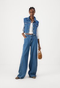 Jeanski brezrokavnik in široki jeans v modri barvi, kombinirani z belim skrajšanim topom. Rjava torbica in čevlji z leopardjim vzorcem. Gladke teksture in sproščen design.