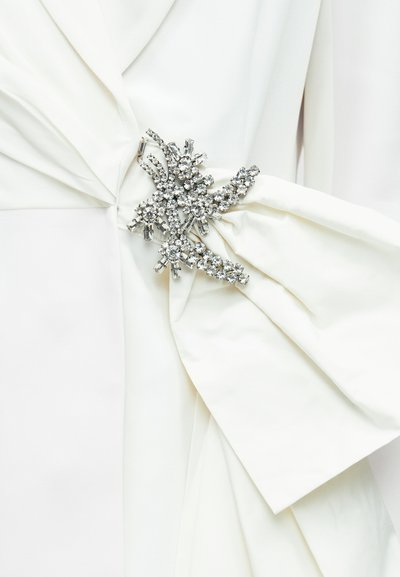 Vestido de tecido branco com pormenor franzido na cintura, adornado com um broche metálico prateado com várias pedras de strass transparentes.