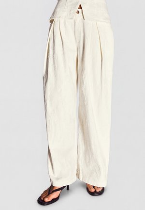 Cremefarbene, hoch taillierte, weit geschnittene Hose mit vorderen Falten, getragen mit schwarzen, zehenfreien Sandaletten mit Absatz vor neutralem Hintergrund.