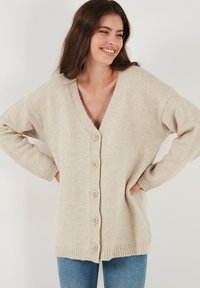 Cardigan beige lavorato a maglia con scollo a V e cinque bottoni, dotato di polsini e orlo a costine. Vestibilità casual e oversize con una texture morbida.