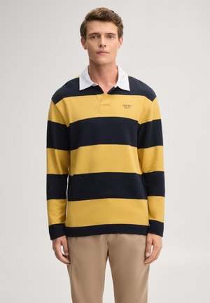 Mann trägt ein langärmeliges Rugbyshirt mit breiten gelben und marineblauen horizontalen Streifen und einem weißen Kragen, steht vor einem einfarbigen Hintergrund.