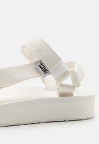 Valkoinen sandaali, jossa on teksturoitu pohja, säädettävät kangasnauhat ja pehmustettu pohja. Teva-logo on kiinnitetty nauhaan.