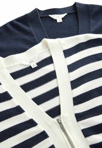 Due maglioni: uno blu navy a tinta unita, l'altro bianco con strisce navy. Entrambi hanno un collo a V e dettagli con zip. Il materiale sembra morbido e testurizzato.