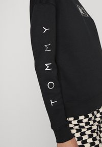 Svart sweatshirtärm med silvermetallisk "TOMMY"-tryck, kombinerad med rutiga svart-vita byxor.
