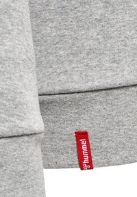 Gros plan du poignet d'un sweat-shirt gris avec une petite étiquette en tissu rouge avec le label hummel en texte blanc.