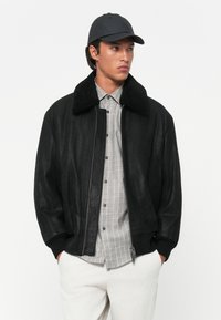 Black leather jacket with a shearling collar, featuring a front zipper, relaxed fit, and worn over a checkered shirt.