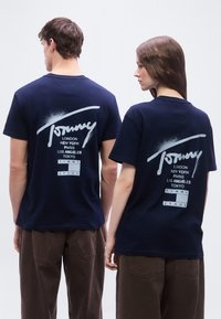 Δύο άτομα που φορούν μπλε ναυτικό T-shirts Tommy Jeans με λευκά ονόματα πόλεων τυπωμένα στην πλάτη, στέκονται δίπλα-δίπλα με την πλάτη στραμμένη προς τα πίσω.