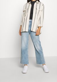 Överdimensionerad skjorta i kräm och svart rand, blå vida jeans och vita sneakers. Har knäppning fram, två bröstfickor och uppvikta ärmar.