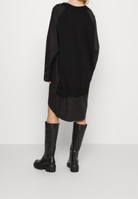 Schwarzes übergroßes Strickkleid mit langen Ärmeln, das einen gerippten Saum und Knopfmanschetten aufweist, kombiniert mit kniehohen schwarzen Lederstiefeln.