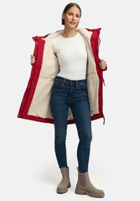 Giacca trapuntata rossa foderata in pile beige, indossata aperta su una camicia bianca aderente e jeans blu. Stivali alla caviglia beige con suole in gomma.