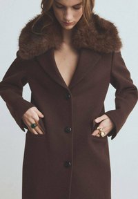 Bruine wollen overjas met een nepbontkraag, voorzien van een afgeslagen revers, zes zwarte knopen en zijzakken. Gladde textuur, getailleerde pasvorm.