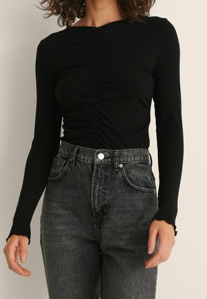 Vrouw draagt een zwarte top met lange mouwen en plooien, ingestopt in een lichtvervaagde zwarte high-waisted spijkerbroek met zichtbare knopen en voorzakken.