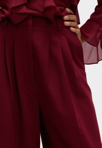 Bordeaux bukser med en skræddersyet pasform, der har plisser, en taljebinding og en glat tekstur. Accenter inkluderer gennemsigtig, rynket stof lagdelt ovenfor.