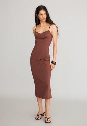 Donna con capelli lunghi e scuri che indossa un vestito midi marrone aderente con spalline sottili e sandali neri con tacco su uno sfondo semplice.