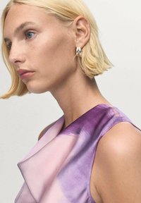 Fioletowo-różowa sukienka bez rękawów o delikatnym dekolcie, gładkiej teksturze i subtelnym połysku. Widać srebrne kolczyki w kształcie kół.