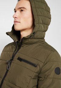 Groene gewatteerde hoodie-jack gemaakt van lichtgewicht materiaal. Bevat een ritszak op de borst en een rond logo op de schouder.