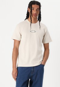Beige cotton t-shirt with short sleeves, featuring a small, gray embroidered logo on the chest. Paired with blue denim jeans.