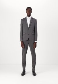 Costume gris au design ajusté, avec un blazer à deux boutons et un pantalon assorti. Chemise blanche en dessous, complétée par des chaussures noires.