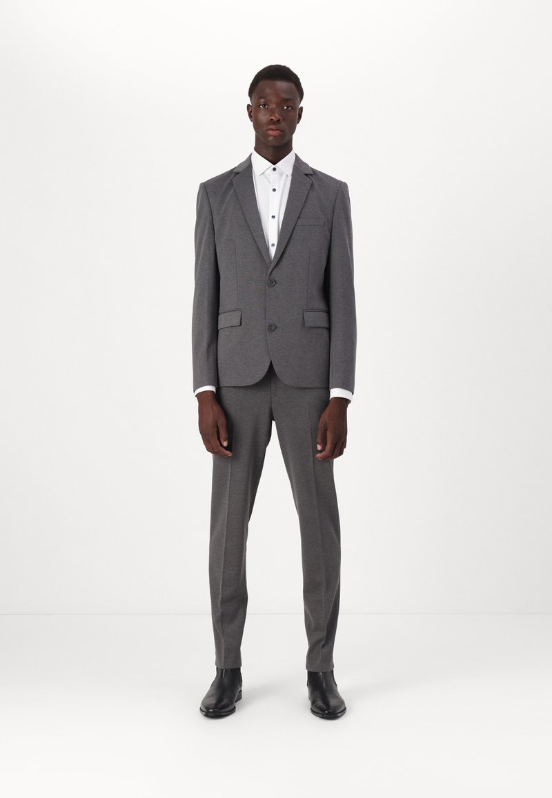 Costume gris au design ajusté, avec un blazer à deux boutons et un pantalon assorti. Chemise blanche en dessous, complétée par des chaussures noires.