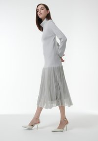 Vrouw die loopt in een lichtgrijs langmouwen jurk met een geplooide rok en witte hoge hakken met enkelbandjes op een witte achtergrond.