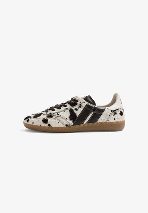 Basket basse avec imprimé vache noir et blanc, rayures marron foncé, lacets noirs et semelle en gomme, vue de côté sur fond blanc.
