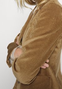 Brun kordfløjl blazer med brede ribber, der har en notch-krave, lange ærmer og to frontlommer. Blød tekstur og varm farve.