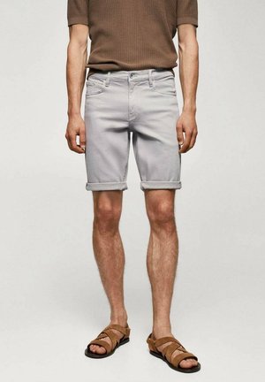 Un homme porte un short gris clair retroussé, un t-shirt en maille à manches courtes marron, et des sandales en cuir beige, se tenant devant un fond uni.