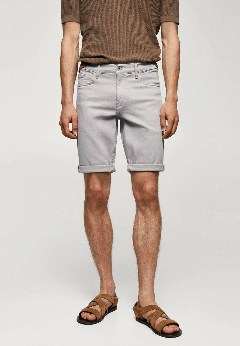 Un homme porte un short gris clair retroussé, un t-shirt en maille à manches courtes marron, et des sandales en cuir beige, se tenant devant un fond uni.
