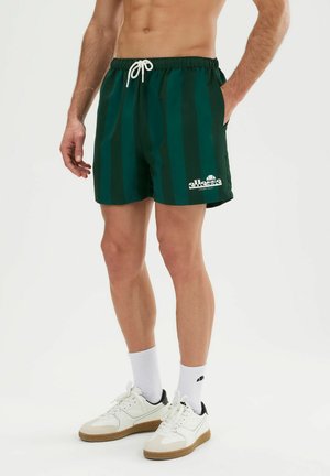 Homme portant un short rayé vert foncé avec un cordon blanc et des baskets blanches avec des chaussettes, debout avec une main dans la poche.