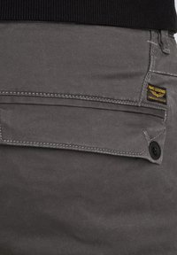 Graue Hose mit glatter Textur, ausgestattet mit einer Klappentasche, kontrastierenden Nähten und einem schwarzen Knopf. Das Etikett weist auf die Marke hin: My Legend.