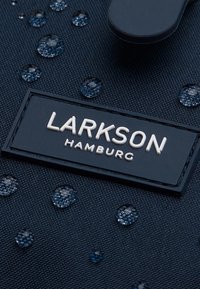 Tejido negro texturizado con gotas de agua, con una etiqueta rectangular que dice "LARKSON HAMBURG" en letras blancas en relieve.