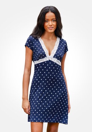Frau trägt ein marineblaues, kurzärmeliges Kleid mit weißen Punkten und weißer Spitzenborte entlang des V-Ausschnitts und der Taille.
